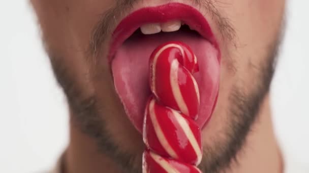 ホットハンサムなひげを生やした男は、唇に赤い口紅を着用し、彼の前に甘い砂糖菓子を保持し、クリックして白い背景のクローズアップで喜びでそれを吸う。女性の裏切りのヒント。正面図. - 映像、動画
