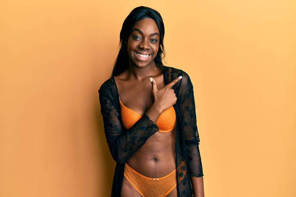 Jovem afro-americana vestindo lingerie alegre com um sorriso de rosto apontando com a mão e o dedo para o lado com expressão feliz e natural no rosto  - Foto, Imagem
