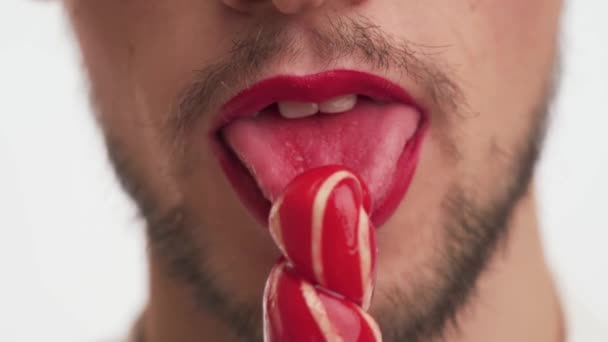 Close-up European attractive fashion man with dark brown beard, moustache wear makeup, smile, blow kiss, hold sweet sugar lollipop, lick, suck it isolated on white background. Mouth view of sexy guy. - Footage, Video