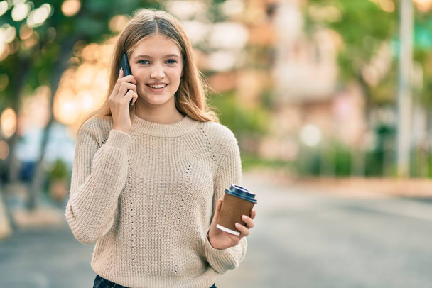 Hermosa adolescente caucásica hablando en el teléfono inteligente bebiendo café en la ciudad. - Foto, imagen