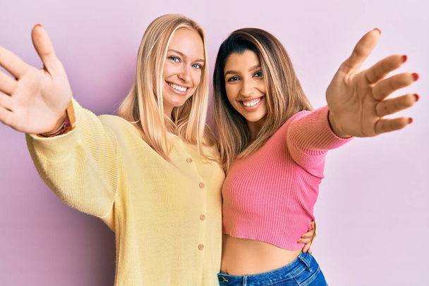 2人の友人が一緒にピンクの背景には、カメラを見て立って抱擁のために腕を開いて笑顔。幸せを受け入れる陽気な表情.  - 写真・画像