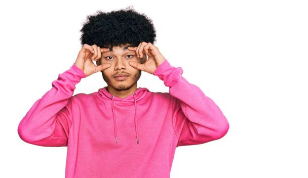 Joven afroamericano con cabello afro usando sudadera rosa casual tratando de abrir los ojos con los dedos, soñoliento y cansado para la fatiga matutina  - Foto, Imagen