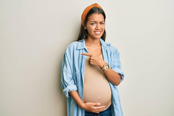 Mulher hispânica bonita esperando um bebê, tocando barriga grávida apontando para o lado preocupado e nervoso com indicador, expressão preocupada e surpreso  - Foto, Imagem