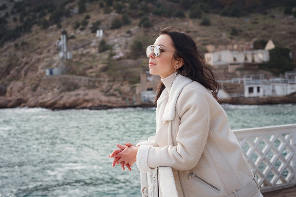 A carefree Caucasian woman in beige clothing enjoying the view of the sea on a warm, windy day. a woman wearing stylish sunglasses enjoying a beautiful winter day and breathing in the sea air - 写真・画像