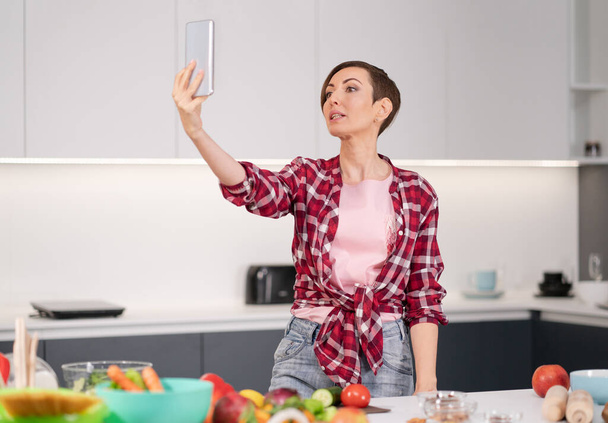 Mulher bonita feliz tirar selfie usando seu smartphone em sua nova casa enquanto cozinha salada fresca vestindo uma camisa xadrez com um estilo de cabelo bob. Partida de comida sã - conceito vegan - Foto, Imagem