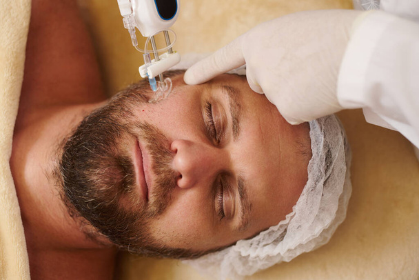 Mãos de esteticista segurando uma arma de mesoterapia e aplicando injeções no rosto de um homem barbudo no salão de spa. - Foto, Imagem