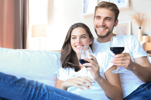 Happy couple sitting, relaxing on couch in living room, using laptop for online shopping together, watching movie, drinking red wine - Zdjęcie, obraz