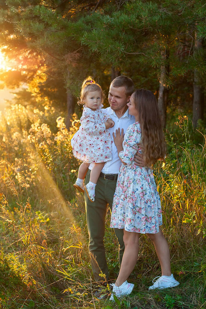 スタイリッシュな若いお母さん、お父さん、娘の家族1歳のブロンドは、夏に木の中の公園の外で、肩の上に父親の近くに座っています。ジーンズの服を着る。家族写真会. - 写真・画像