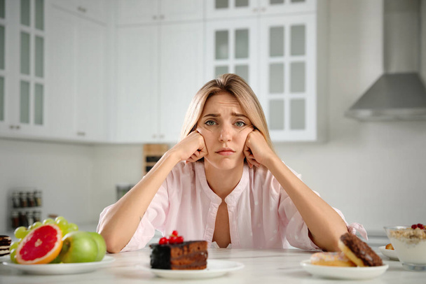 Woman choosing between sweets and healthy food at white table in kitchen - Foto, afbeelding