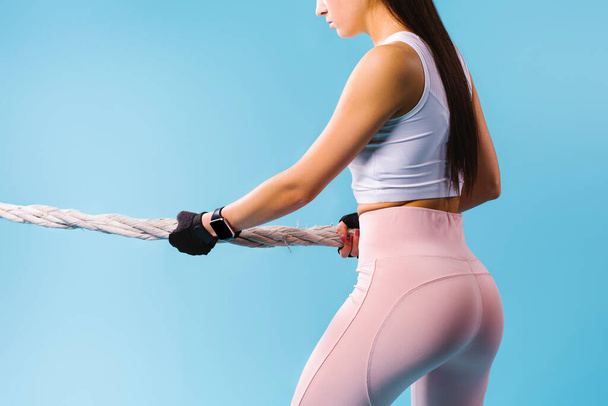 Cropped photo of a girl with pumped up buttocks pulling a rope on a blue background. - Foto, afbeelding