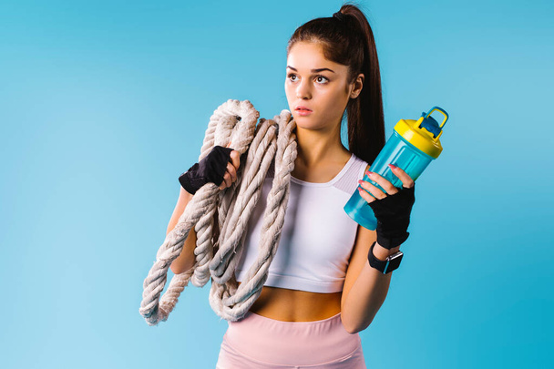 Una chica deportiva con una cuerda viscosa en el hombro y un cóctel de proteínas en la mano mira a un lado sobre un fondo azul con espacio publicitario lateral vacío. - Foto, imagen