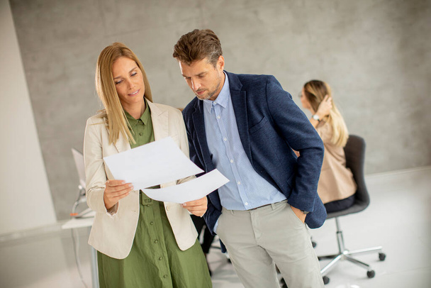 Homme et femme discutant avec du papier dans les mains à l'intérieur du bureau avec des jeunes travaille derrière eux - Photo, image