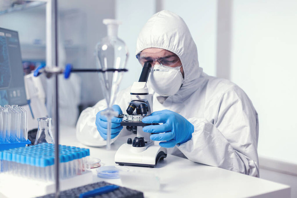 Ученый смотрит в микроскоп, одетый в костюм ppe - Фото, изображение