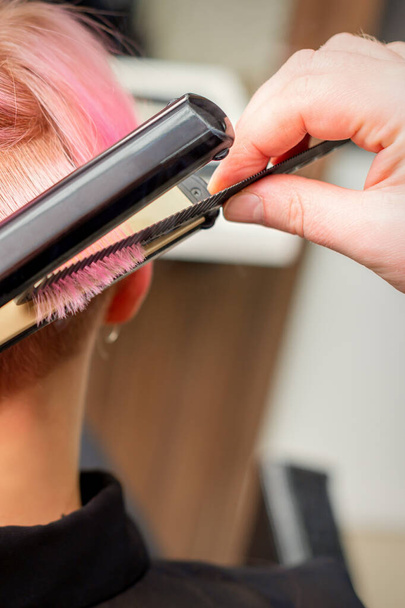 Közelkép a fodrász kiegyenesítése a rövid rózsaszín haj egy női ügyfél egy hajkiegyenesítő vas egy szépségszalonban - Fotó, kép