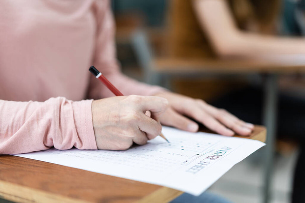 Κοντινό πλάνο των νεαρών γυναικών φοιτητών πανεπιστημίου επικεντρωθεί στην πραγματοποίηση εξετάσεων στην τάξη. Κορίτσι φοιτητής γράφει την απάντηση των εξετάσεων στο φύλλο απάντησης στην τάξη. - Φωτογραφία, εικόνα