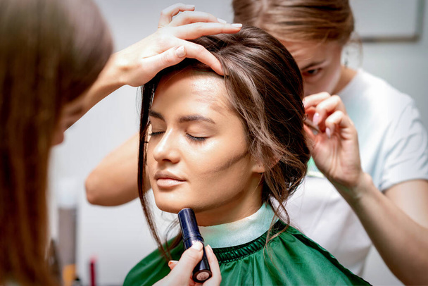 De kapper en make-up kunstenaar doen kapsel en make-up voor een mooie jonge blanke vrouw in een schoonheidssalon - Foto, afbeelding