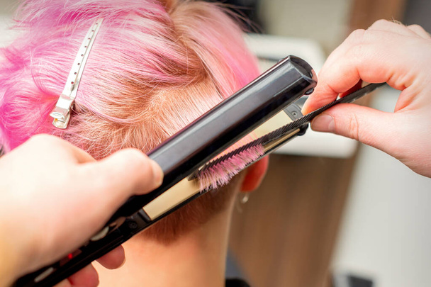 Gros plan du coiffeur redressant les cheveux roses courts d'une cliente avec un fer à lisser dans un salon de beauté - Photo, image