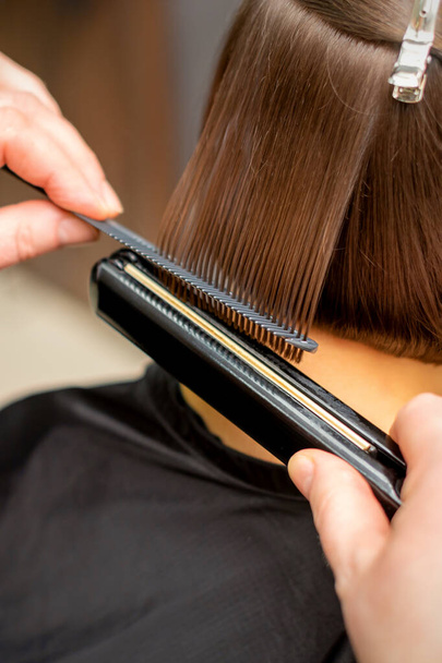 Close up of the hairdresser straightening the short hair of a female client with a hair straightening iron in a beauty salon - Foto, afbeelding