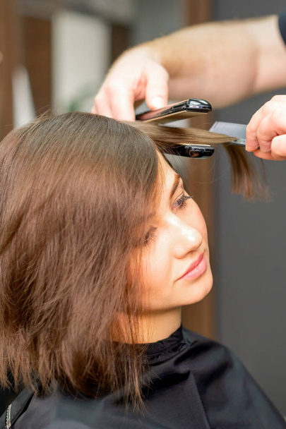 Un parrucchiere maschile sta raddrizzando i capelli della giovane donna in un salone di bellezza - Foto, immagini