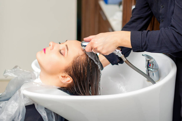 Una peluquera está enjuagando el cabello de una joven en un lavabo después de lavarse el cabello en una peluquería - Foto, imagen