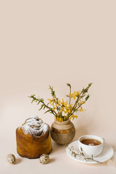 Easter cake craft in. Craft with raisins, candied fruits and a sprinkle of powdered sugar. Homemade cake. Eggs, a cup of tea and spring flowers in a vase on a beige background - Foto, Bild