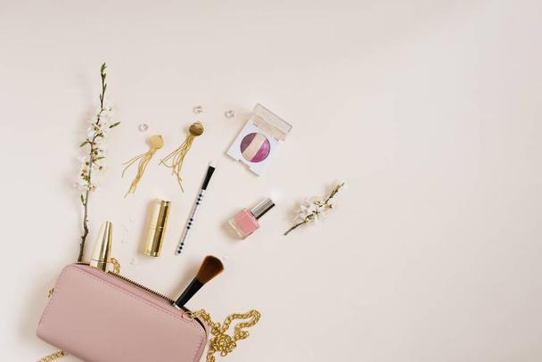 Női asztal kozmetikumokkal, amely egy rózsaszín táska vagy kozmetikai táska mellett található, fénymásoló hellyel, almafa virágokkal bézs alapon. Első látásra. Lapos volt. A keret összetétele anyák napjára vagy bloggerre. - Fotó, kép