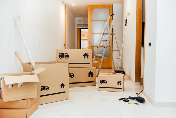 Pile of Cardboard boxes being opened after moving in empty room, house, apartment. Home renovation, moving or relocation concept. Repairing equipment, Drilling machine, paint roller. No people. - Photo, Image