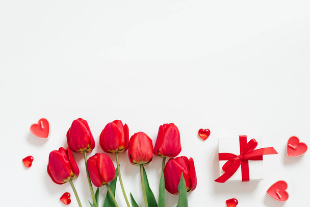 Valentijnsdag Romantische achtergrond. Rode tulpen, een geschenk met een strik, en kaarsharten op een witte achtergrond met kopieerruimte. Vlakke plaat - Foto, afbeelding