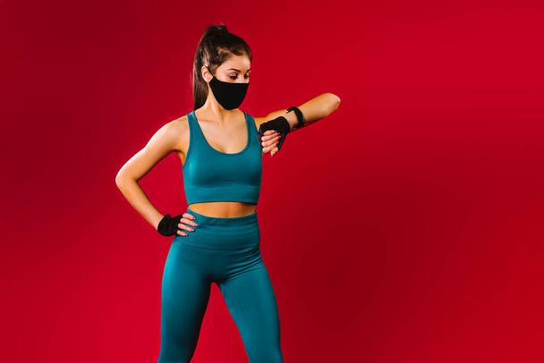 Caucasian fitness girl wearing a protective mask looks at her achievements on a fitness watch during training. Sports during quarantine. Red background and side advertising space. - Photo, Image