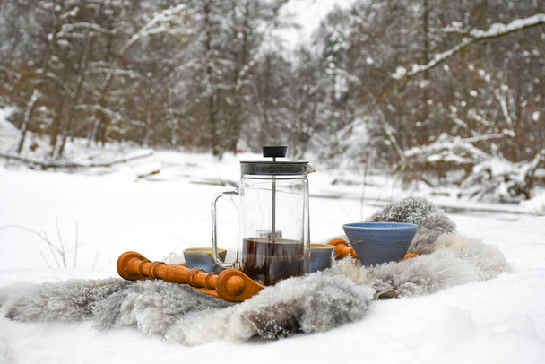 Drinking coffee or tea on the snow in winter with cups, warm blankets and winter landscape with river and forest on background - Photo, Image