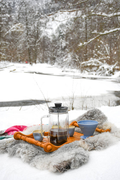 Drinking coffee or tea on the snow in winter with cups, warm blankets and winter landscape with river and forest on background, vertical  - Photo, Image