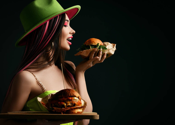 Beautiful woman hold two craft big burger sandwiches with hungry open mouth ready to eat in green popular hat happy on black  - 写真・画像