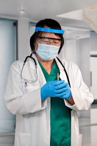 Medico donna asiatica con scudo facciale e guanti che tiene una siringa con entrambe le mani. Concentrazione selettiva. Concetto di vaccinazione e assistenza sanitaria. - Foto, immagini