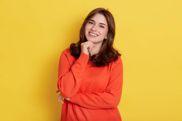 European charming girl with dark hair, holding fist under chin and smiling broadly, looking directly at camera, posing isolated over yellow background, expressing happiness. - Фото, зображення