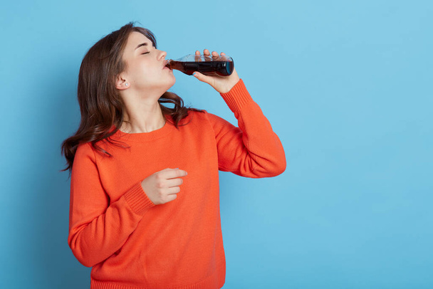 Thirsty female wearing orange jumper standing isolated over blue background, holding bottle of fizzy water and drinking, adorable girl drinks sweet beverage, enjoying tasty soda. - Photo, image