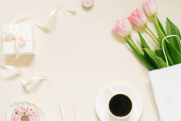 Poranna filiżanka kawy, pączka, prezent lub pudełko i wiosenne kwiaty tulipanów na beżowym tle. Piękne śniadanie dla kobiet, Dzień matki, Walentynki. Układy płaskie. - Zdjęcie, obraz