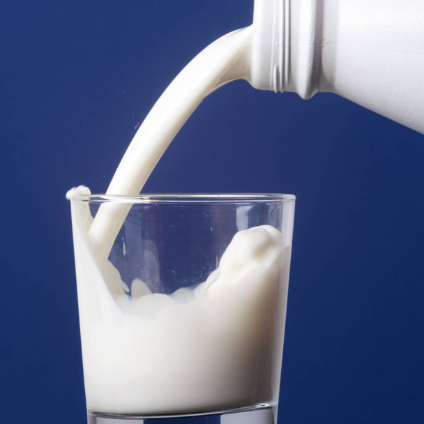 Verter la leche de la botella de plástico en un vaso sobre fondo azul - Foto, Imagen