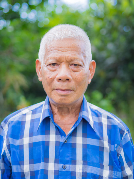 Retrato de homem idoso vestindo uma camisa azul de pé, sorrindo e olhando para a câmera em seu jardim. Fundo de natureza verde bonita - Foto, Imagem