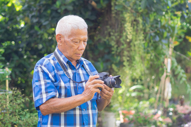 Senior man maakt foto 's met digitale camera in de tuin. Oude man draagt een blauw shirt, blij als hij een camera gebruikt. Fotografieconcept - Foto, afbeelding