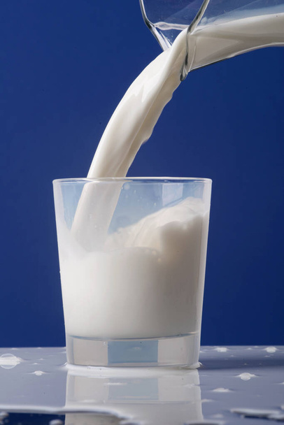 Ρίχνουμε το κρεμώδες γάλα από την καράφα ή την κανάτα σε ένα ποτήρι. Το γάλα χύνεται πάνω στο τραπέζι. Φωτογραφία απομονωμένη σε μπλε φόντο - Φωτογραφία, εικόνα