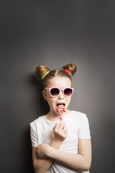 Bright colors: a girl with a striped red lollipop and sunglasses on a gray background. - Foto, imagen