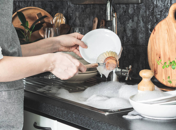 Mulher incerta no avental cinza lava pratos com a escova de madeira perto da pia na cozinha. Vista frontal - Foto, Imagem