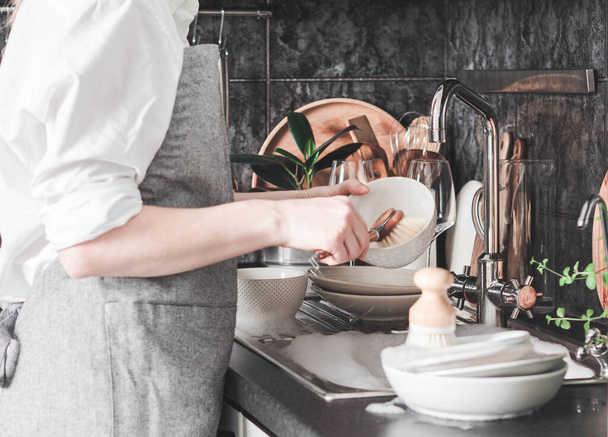 Mulher incerta no avental cinza lava pratos com a escova de madeira perto da pia na cozinha. Vista frontal - Foto, Imagem