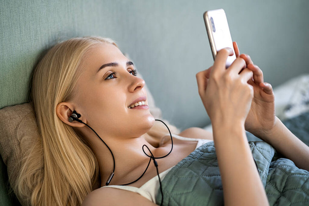 Smiling girl listening music on her smartphone lying on bed in a bedroom - Foto, Imagem
