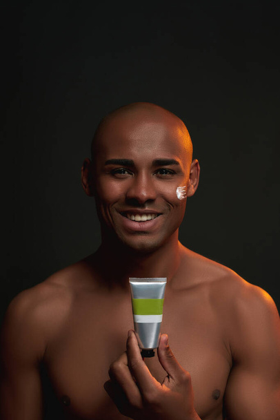 Portrait of shirtless african american man smiling at camera, holding beauty product while posing with cream applied on his cheek isolated over black background - Foto, imagen
