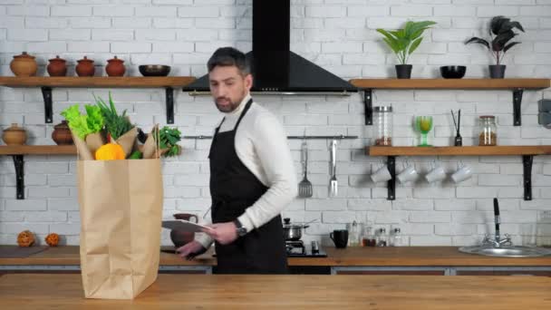 Hombre de delantal negro pone cuchillo y tabla para cortar verduras en la mesa de la cocina - Imágenes, Vídeo