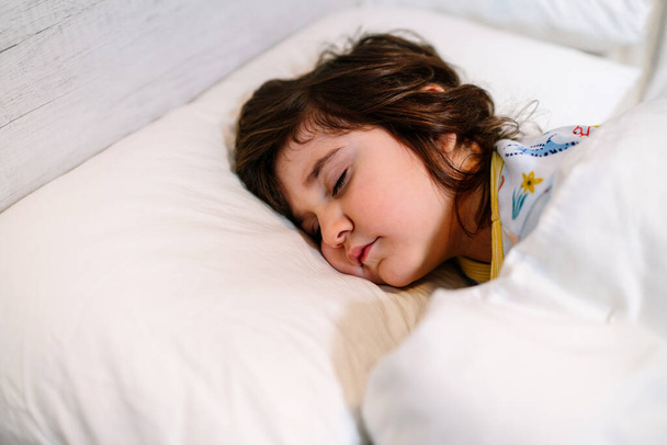 child asleep in bed with closed eyes and innocent face, wearing pajamas and white sheets - Photo, Image