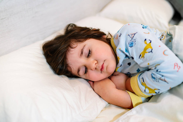niño dormido en la cama con los ojos cerrados y la cara inocente, usando pijamas y sábanas blancas - Foto, Imagen