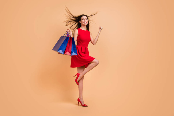 Full size profile side photo of brunette nice girl raise fists win hold bags fly hair wear red high-heels isolated on beige color background - Foto, afbeelding