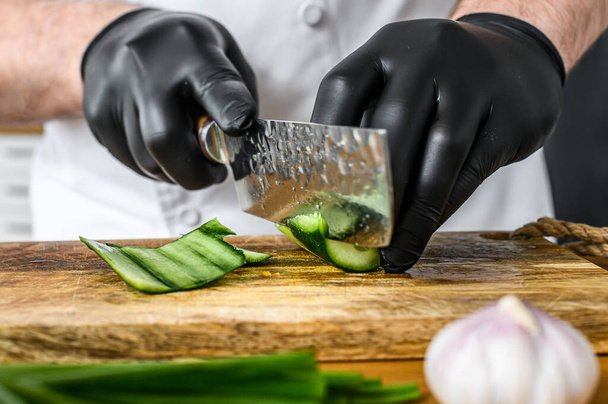 Un chef con guantes negros está cortando un pepino verde fresco en una tabla de cortar de madera. Concepto de cocinar alimentos orgánicos saludables - Foto, Imagen
